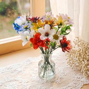Puede incluir: Un colorido ramo de flores de cristal en un peque&ntilde;o jarr&oacute;n de cristal transparente. Las flores son de varios colores, incluyendo rojo, blanco, amarillo, azul y morado. El jarr&oacute;n est&aacute; sobre un mantel de encaje blanco cerca de una ventana.