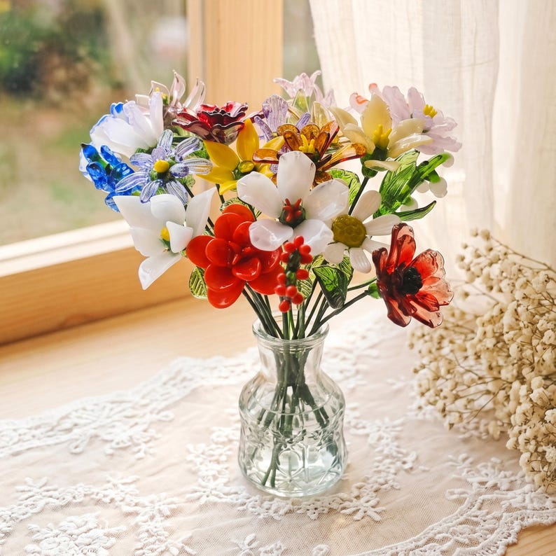 Puede incluir: Un colorido ramo de flores de cristal en un peque&ntilde;o jarr&oacute;n de cristal transparente. Las flores son de varios colores, incluyendo rojo, blanco, amarillo, azul y morado. El jarr&oacute;n est&aacute; sobre un mantel de encaje blanco cerca de una ventana.