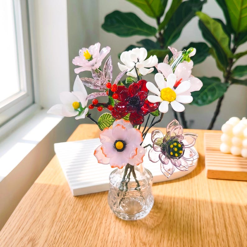 Puede incluir: Un jarr&oacute;n de cristal contiene un colorido ramo de flores artificiales. El arreglo incluye margaritas blancas, flores rosas y rojas y hojas verdes. El jarr&oacute;n est&aacute; sobre una mesa de madera cerca de una ventana.