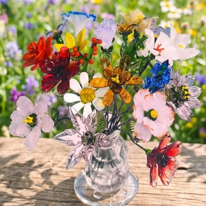 Puede incluir: Un colorido ramo de flores de cristal en un jarr&oacute;n transparente. El arreglo incluye flores rojas, naranjas, amarillas, blancas, rosas, azules y moradas con tallos verdes. El jarr&oacute;n est&aacute; sobre una superficie de madera.