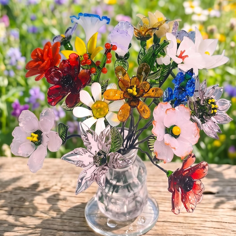 Puede incluir: Un colorido ramo de flores de cristal en un jarr&oacute;n transparente. El arreglo incluye flores rojas, naranjas, amarillas, blancas, rosas, azules y moradas con tallos verdes. El jarr&oacute;n est&aacute; sobre una superficie de madera.