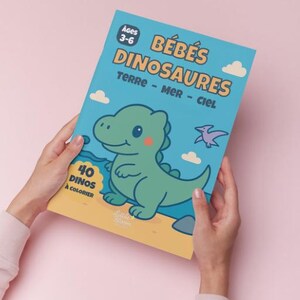 May include: A children's coloring book titled "B&eacute;b&eacute;s Dinosaures" held against a pink background. The cover is light blue with a cartoon dinosaur illustration. Text includes "Ages 3-6", "40 Dinos &agrave; Colorier", and "Terre - Mer - Ciel".