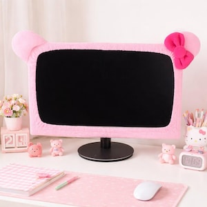 May include: A pink computer monitor cover with a cartoon-style design, featuring ears and a bow. The cover is on a black monitor, surrounded by pink desk accessories, including a mouse, notepad, and small figurines.