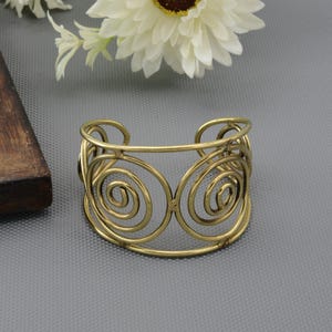 Op de afbeelding: Een goudkleurige metalen manchetarmband met een opengewerkt ontwerp bestaande uit meerdere onderling verbonden draadspiralen. De armband heeft een brede, cirkelvormige structuur met een gepolijste afwerking en een open achterkant.