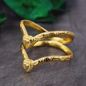 May include: A gold-colored ring with a double-band design. The ring features two heart-shaped accents at the ends of the bands. The bands have a textured, engraved pattern. The ring is displayed on a dark gray surface.