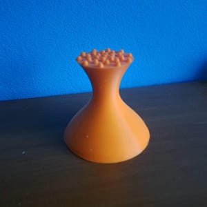 Könnte beinhalten: Ein orangefarbenes, sanduhrförmiges Objekt mit breiter Basis und einem ausgestellten Oberteil, das mit kleinen, abgerundeten Noppen bedeckt ist. Das Objekt besteht aus einem festen Material und steht auf einer dunkelbraunen Oberfläche vor einem blauen Hintergrund.