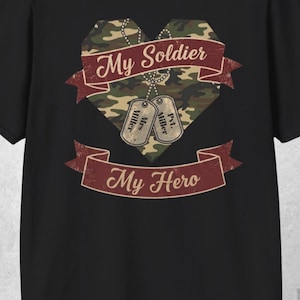 Könnte beinhalten: Schwarzes T-Shirt mit einem Tarnmuster-Herz-Design und den Worten "My Soldier" und "My Hero" auf weinroten Bannern. Enthält zwei Erkennungsmarken mit dem Namen "Miller".