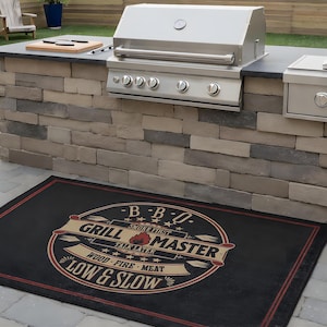Pode incluir: Grelhador de exterior em aço inoxidável com queimador lateral embutido e tapete preto. O tapete apresenta o texto "GRILL MASTER" num design circular com as palavras "LOW & SLOW". O grelhador está sobre uma estrutura de pedra.