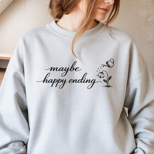 May include: A pale grey sweatshirt featuring the phrase "maybe happy ending" in black cursive script. A small black illustration of a watering can watering a flower is to the right. The sweatshirt has a crew neck.