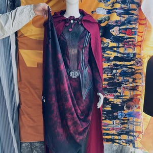 Pode incluir: Um conjunto de fantasia composto por uma capa com capuz vermelho-escura com bainha desgastada, uma túnica a condizer com padrão vermelho-escuro e preto com cinto central e um toucado vermelho-escuro num manequim com fundo amarelo.