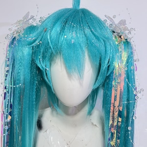 May include: A bright teal wig styled with two long pigtails and a small top tuft. The hair features iridescent tinsel strands, clear beaded wire accents, and small white butterfly embellishments attached to the sides of the pigtails.