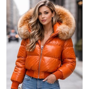 May include: An orange puffer jacket with a fur-lined hood. The jacket has a shiny, glossy finish and a ribbed hem and cuffs. The model is wearing blue jeans. The jacket is a stylish outerwear option for cold weather.