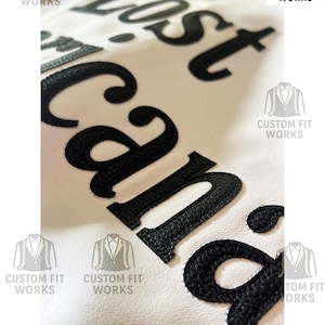 May include: Close-up of a white leather jacket with the words "Lost Cana" embroidered in black. The jacket features the "Custom Fit Works" logo in multiple locations.