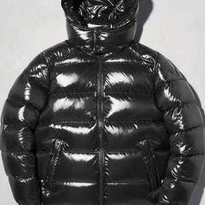 May include: A shiny black puffer jacket with a hood. The jacket features a full-length zip, two side pockets, and a quilted pattern. The glossy material reflects light, giving it a sleek, modern appearance. This winter coat is designed for warmth and style.