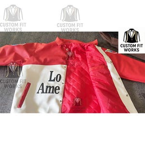 May include: A red and white jacket with the text "Lo Ame" on the front. The jacket has a quilted red interior and white stripes along the sleeves. The jacket has the text "CUSTOM FIT WORKS" on it.