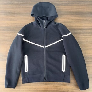 Puede incluir: Chaqueta negra con capucha y cremallera, con rayas blancas en el pecho y los brazos. La chaqueta tiene capucha, dos bolsillos delanteros y una cremallera completa. El material es liso.