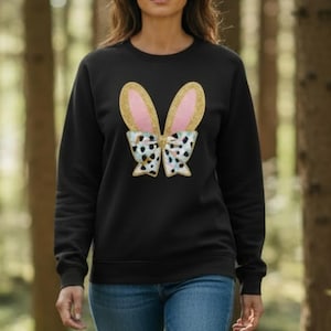 May include: Black sweatshirt with a bunny ears design. The ears are gold glitter with pink inner ears, and the bow tie is white with a black and tan spotted pattern. A casual top for Easter or spring.