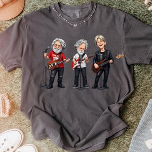 May include: Dark gray t-shirt featuring a cartoon illustration of three musicians playing guitars and singing into microphones. The musicians are dressed in casual attire. The shirt has a crew neck and short sleeves.