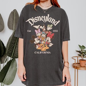 Op de afbeelding: Donkergrijs T-shirt met het Disneyland-logo en de stripfiguren Mickey Mouse, Minnie Mouse, Donald Duck, Daisy Duck, Goofy en Pluto. Het shirt bevat de tekst "Est 1955" en "California".