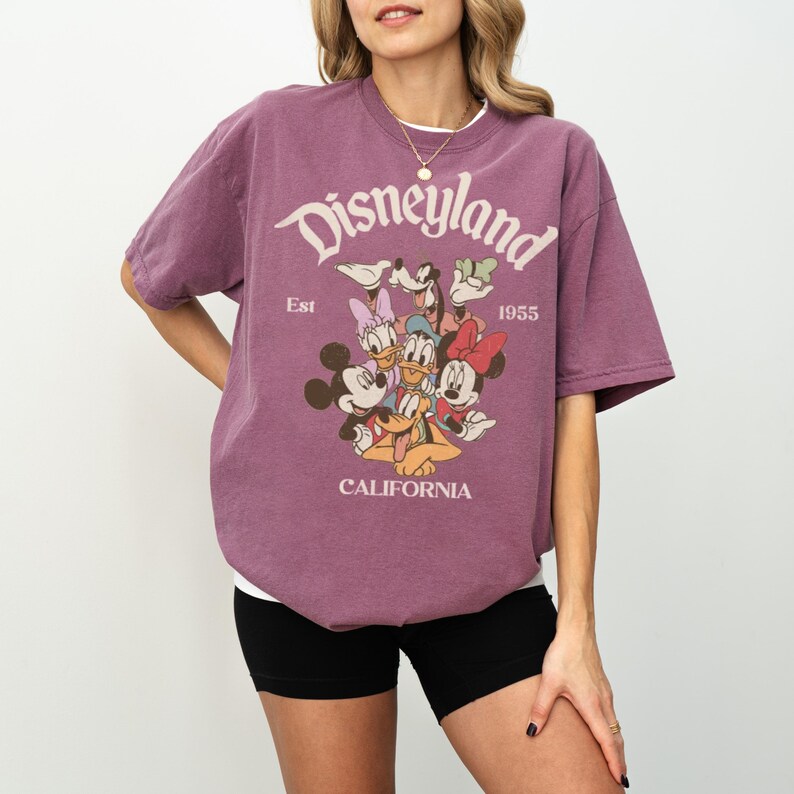 Op de afbeelding: Een mauvekleurig T-shirt met het Disneyland-logo en een afbeelding van Disney-personages. De tekst "Disneyland Est 1955 California" is op de shirt gedrukt. Het shirt wordt gedragen met een zwarte short.