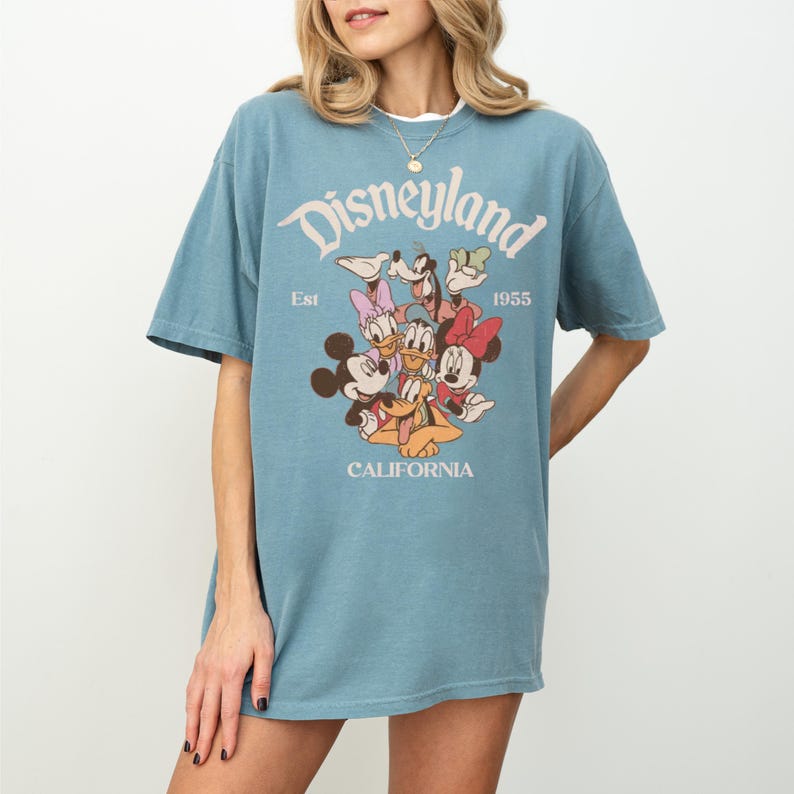Op de afbeelding: Een blauw t-shirt met het woord "Disneyland" in een vintage lettertype, de tekst "Est 1955" en "California". Het shirt heeft een afbeelding van Mickey Mouse, Minnie Mouse, Donald Duck, Daisy Duck, Goofy en Pluto.