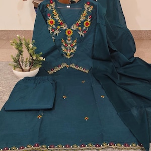May include: Teal embroidered salwar kameez set. The top has a V-neck with red, yellow, and green floral embroidery. The set includes matching trousers and a dupatta. The hemline and sleeves have a decorative border.