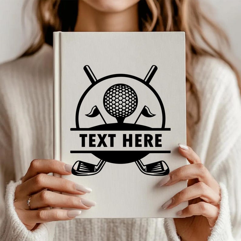 May include: A white book cover featuring a black golf-themed design. The design includes a golf ball, flags, golf clubs, and the text "TEXT HERE" in a bold font. The book is held by a person.