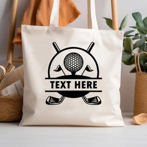 May include: Cream-colored tote bag featuring a black golf-themed design. The design includes a golf ball on a tee, flags, golf clubs, and the text "TEXT HERE" in a horizontal banner. The bag has two handles.