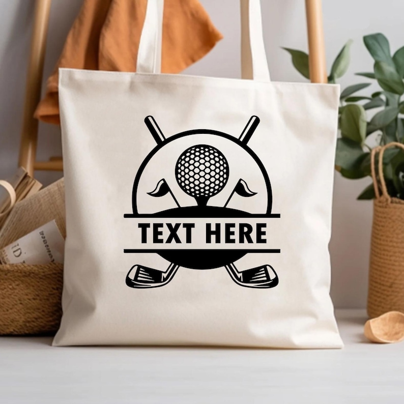 May include: Cream-colored tote bag featuring a black golf-themed design. The design includes a golf ball on a tee, flags, golf clubs, and the text "TEXT HERE" in a horizontal banner. The bag has two handles.