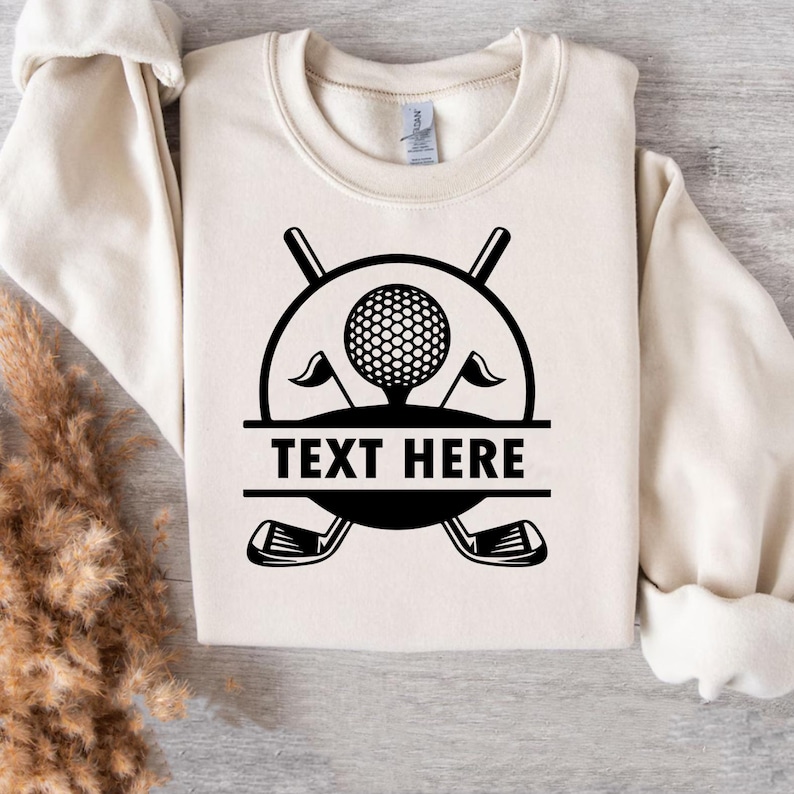 May include: A cream-colored sweatshirt with a black golf-themed graphic. The graphic includes a golf ball, flags, crossed golf clubs, and the text "TEXT HERE" in a bold font. The sweatshirt is folded.