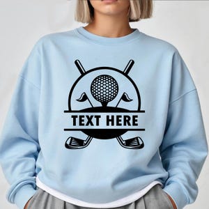 May include: Light blue sweatshirt featuring a black golf-themed graphic. The design includes a golf ball, crossed golf clubs, flags, and the text "TEXT HERE" in a bold font. The sweatshirt has a crew neck and long sleeves.