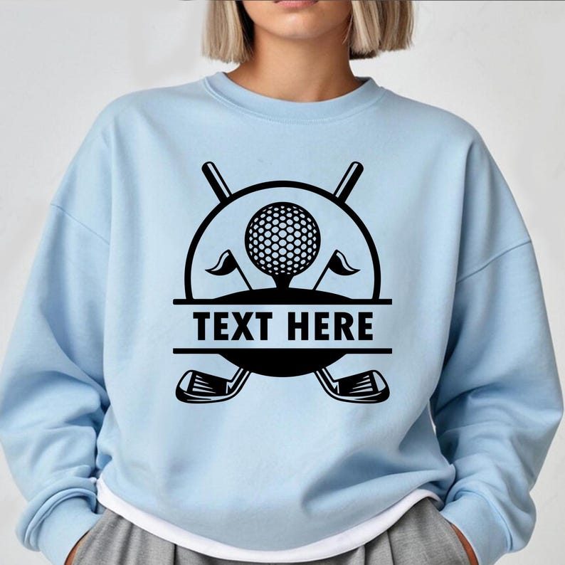 May include: Light blue sweatshirt featuring a black golf-themed graphic. The design includes a golf ball, crossed golf clubs, flags, and the text "TEXT HERE" in a bold font. The sweatshirt has a crew neck and long sleeves.