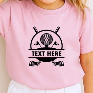 May include: Pink t-shirt featuring a black golf-themed graphic. The design includes a golf ball, flags, golf clubs, and the text "TEXT HERE" in a rectangular box. The shirt is made of a soft material.