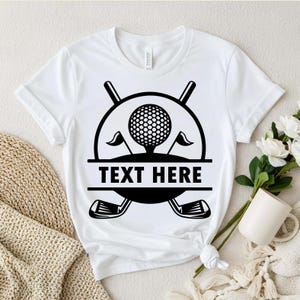 May include: White t-shirt featuring a black graphic of a golf ball, flags, and clubs. The design includes the text "TEXT HERE" in a horizontal bar. The shirt is a crew neck style.