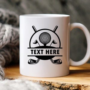 May include: White ceramic mug featuring a black golf-themed design. The design includes a golf ball, flags, golf clubs, and the text "TEXT HERE" in a bold font. The mug has a curved handle.