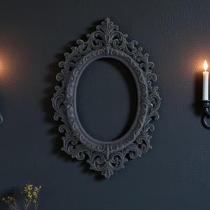 Può includere: Una cornice ovale grigio scuro con dettagli ornati è appesa a una parete scura, affiancata da due candelieri con candele accese. Un piccolo vaso con fiori secchi, una rivista e una brocca di ceramica bianca sono su un tavolo di legno.