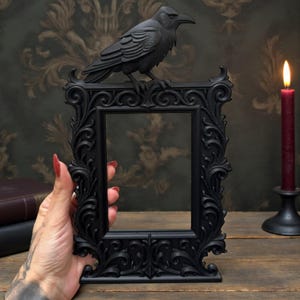 May include: A black, decorative picture frame with a raven figurine. The frame has an elaborate, swirling design and is held by a person. A lit red candle and two leather-bound books are in the background, with a patterned wallpaper.
