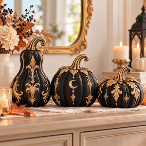 Può includere: Tre zucche decorative in nero opaco con filigrana e foglie in oro metallizzato. La più alta è a forma di pera, quella centrale presenta una mezzaluna e la più piccola è rotonda. Ognuna ha un gambo dorato ricurvo e dettagli intricati.