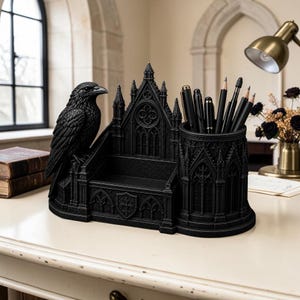 Könnte beinhalten: Ein schwarzer Schreibtisch-Organizer mit einem skulptierten Raben auf der linken Seite, einem zentralen Fach und einem zylindrischen Stiftehalter auf der rechten Seite. Die Struktur ist mit Spitzbögen und architektonischen Motiven verziert.
