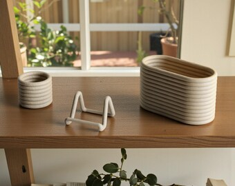 May include: A set of three off-white objects consisting of a small cylindrical container with ribbed walls, a larger oval-shaped container with matching ribbed walls, and a small, angular, four-legged stand positioned between the two containers.