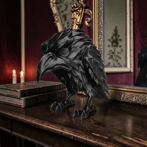 May include: A matte black raven figurine featuring detailed, layered feather textures across its wings and body, a sharp curved beak, and prominent talons. The bird is depicted in a standing position with its head slightly bowed forward.