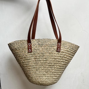 May include: A woven straw tote bag with brown leather handles. The bag has a wide, open top and a rounded bottom. The handles are attached to the bag with small leather tabs. The bag is held up by a hand.