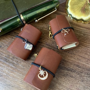 Op de afbeelding: Drie kleine, bruine leren notitieboekjes met zwarte elastiekjes en gouden bedels. Eén bedel is een huis, een andere een strik en de derde een maan en ster. Een groene pen en een gouden ginkgoblad staan op de achtergrond.