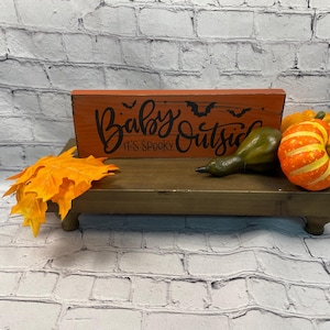 May include: A wooden riser with a decorative orange sign that reads "Baby It's Spooky Outside" with bat silhouettes. The riser also holds a decorative gourd, a small orange and yellow pumpkin, and orange fall leaves.