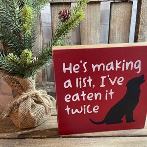 May include: Red wooden sign with white text that reads "He's making a list, I've eaten it twice". A black silhouette of a dog is on the right side of the sign.