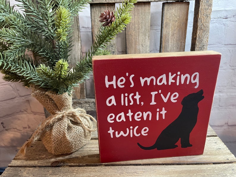May include: Red wooden sign with white text that reads "He's making a list, I've eaten it twice". A black silhouette of a dog is on the right side of the sign.