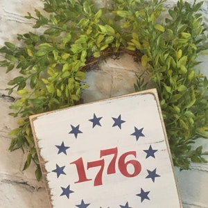 May include: A white wooden sign with a red "1776" surrounded by a circle of blue stars. The sign is distressed and has a rustic look.