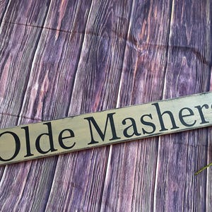 May include: A rustic wooden sign with the text "Olde Mashers" painted in black lettering on a light brown background.