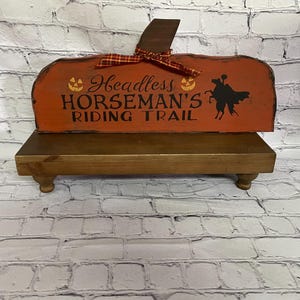 May include: A wooden pumpkin-shaped sign with the text "Headless Horseman's Riding Trail" in black. The sign is orange with a brown stem and a plaid ribbon. It sits on a small wooden stand with rounded legs, perfect for Halloween decor.