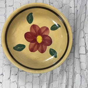May include: A round, yellow ceramic bowl with a floral design. The bowl features a large red flower with a yellow center and green leaves. A dark brown band encircles the rim.